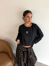 Remera de manga larga color negro, con cuello redondo y corte cropped. Presenta un pequeño parche cuadrado con diseño gráfico blanco en el centro del pecho.