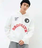 Hoodie blanco con capucha y cordones ajustables, confeccionado en algodón con felpa interior. Presenta estampado frontal con la palabra 'BROOKLYN' en color rojo, un parche circular con la imagen de Notorious B.I.G. y una firma bordada. Cuenta con bolsillo canguro y puños ajustados.