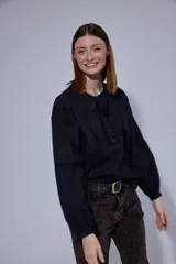 Camisa blanca de algodón con pechera decorada con alforzas y puntillas. Presenta cierre frontal con botones, mangas tres cuartos con frunces y detalles de puntilla, y un diseño de corte holgado con ruedo redondeado.