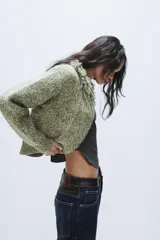 Chaqueta de punto de textura jaspeada en color verde, con cuello redondo adornado con flecos del mismo tejido, manga larga y cierre frontal con corchete metálico.