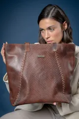 Bolso tipo shopper de cuero marrón con textura tejida y detalles de costuras decorativas en los laterales. Cuenta con doble asa de hombro, cierre principal y placa metálica con el logo de la marca Pecarí en el frente.