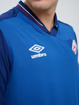 Remera polo deportiva Umbro del Club Nacional de Football, color azul con cuello en V y detalles en rojo y blanco. Incluye el logo de Umbro y el escudo del club.