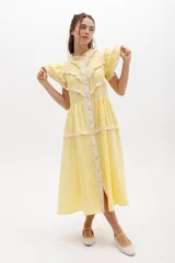 Vestido midi amarillo claro de lino, con cuello mao, cierre frontal de botones y volados en las mangas. Presenta detalles de volados y ribetes en color beige a lo largo de la botonera y en los bordes de los volados del cuerpo y falda.