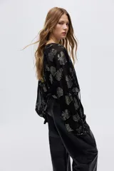 Blusa de corte holgado en tejido jacquard transparente con diseño de hojas metalizadas. Presenta cuello mao con frunces, abertura frontal con tiras finas, ribete de volados y mangas largas tipo globo con puños elásticos.