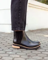 Botinetas tipo Chelsea de cuero negro, con paneles laterales elásticos y detalle de textura grabada en el talón. Presentan una suela plana con plataforma de madera clara y base de goma negra.