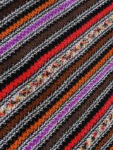 Capa o poncho de tejido de punto con cuello en V y flecos en el borde inferior. Presenta un patrón de rayas verticales en zigzag con una rica paleta de colores que incluye tonos marrones, negros, rojos, naranjas y morados.