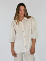 Camisa de algodón a rayas beige y blancas, con cuello camisero, botones delanteros naranjas y mangas abullonadas de tres cuartos.