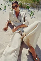 Pantalón ancho de tiro alto color beige claro, con cintura elástica, cordón ajustable y pinzas delanteras. Presenta bolsillos frontales y bolsillos de plastrón en la espalda. Incluye un cinturón del mismo tejido con apliques metálicos.