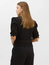 Blusa de algodón semitransparente con diseño floral bordado a tono. Presenta escote en V, cierre frontal con botones y mangas al codo ligeramente abullonadas. Incluye detalles de encaje calado estilo macramé en los puños y en el ruedo con terminaciones en ondas.