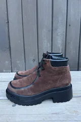 Botas de caña corta confeccionadas en cuero nobuk color marrón, con detalles en cuero negro en los bordes y cordones negros. Presentan una suela de plataforma dentada de color negro.