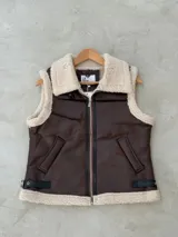 Chaleco de cuero sintético en color marrón con interior y bordes de corderito sintético color crema. Presenta cierre frontal, bolsillos laterales y detalles de correas ajustables con hebillas en la parte inferior.