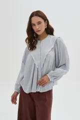 Blusa de algodón color beige, con cuello redondo y cierre frontal con botones. Presenta delicados bordados decorativos en tono tostado sobre el pecho y los hombros, mangas largas abullonadas con puños ajustados y diseño holgado.