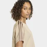 Remera Adidas Firebird de corte holgado, confeccionada en tejido tricot color beige. Presenta cuello redondo, mangas cortas con las icónicas tres rayas en color marrón y el logo del trébol bordado en el pecho.