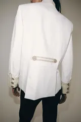 Chaqueta de estilo militar de ZARA Woman Collection, confeccionada en mezcla de algodón en color blanco y beige. Presenta cuello subido, manga larga con puños anchos en contraste y cierre frontal con alamares y botones metálicos en relieve. Tiene bajo asimétrico con abertura en la espalda.