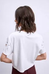 Remera blanca de algodón con cuello redondo y mangas cortas. Presenta apliques metálicos y aberturas circulares en los hombros y el frente.