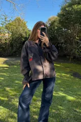 Camisa oversize a rayas marrón y negro, con cuello camisero, bolsillo frontal grande con bordado rojo de un caballo, y mangas abullonadas con puños elásticos.