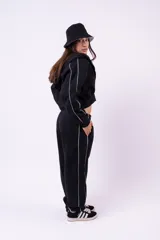Conjunto deportivo compuesto por una campera con cierre y un pantalón de jogging, ambos de color negro con detalles de líneas blancas laterales. El pantalón presenta un corte holgado y cómodo.