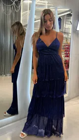 Vestido largo de fiesta color azul marino, con acabado brillante y textura plisada. Presenta escote en V, breteles finos y falda con tres niveles de volados.