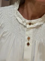 Camisa de algodón color blanco con cuello redondo y detalle de fruncido tipo nido de abeja en el canesú. Presenta cierre frontal con botones decorativos, mangas largas abullonadas con puños elásticos fruncidos y volados en el cuello.