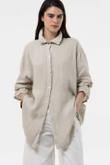 Camisa de lino color beige, de calce amplio y relajado, con cuello camisero, cierre frontal de botones, mangas largas y diseño de largo irregular con aberturas laterales.