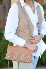 Cartera tipo sobre de cuero color camel con detalle de tachas metálicas plateadas en la parte frontal. Incluye una correa de cadena metálica plateada y cierre superior.