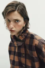 Camisa de manga larga con estampado de cuadros en tonos marrón y negro. Presenta un diseño cruzado con cuello vuelto y cierre frontal de botones, con un corte asimétrico en el bajo.