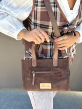 Cartera bandolera de cuero color marrón chocolate, con forro de tela y herrajes dorados. Tiene doble asa de mano y correa larga regulable y desmontable. Incluye bolsillo frontal con cierre y bolsillo interno.