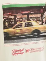 Camiseta de manga corta y cuello redondo, color blanco antiguo (off-white), con un estampado gráfico grande en la espalda que simula una fotografía urbana nocturna con un taxi amarillo y texto en tipografía cursiva y recta.