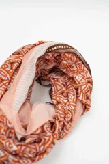 Pañuelo rectangular de tejido fino con estampado geométrico y tribal en tonos naranja, terracota y rosa pálido, con bordes en rayas verticales.