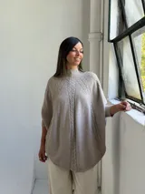Poncho de tejido de punto color visón, con cuello alto y diseño de trenzas verticales. Presenta mangas cortas ajustadas y un corte holgado y redondeado en el ruedo.