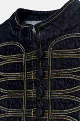 Chaleco de denim azul oscuro con cuello redondo y cierre frontal de botones. Presenta un diseño de pespuntes decorativos en contraste con forma de alamares en la parte delantera.