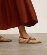 Zapatilla tipo bailarina con capellada tejida con efecto rafia color beige, punta redonda, tira fina marrón con hebilla y suela marrón.