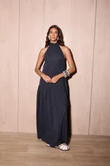 Vestido largo negro, holgado, con escote halter y espalda baja. Presenta un detalle de volado en el cuello y un maxi volado en la cadera. El material es tela Cupro, con apariencia similar a la gamuza.