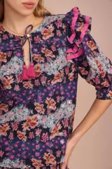 Blusa de algodón con estampado floral en tonos azul, rosa y naranja. Tiene cuello redondo con lazo y mangas cortas abullonadas con volados rosas.