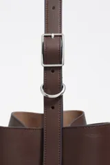 Bolso de hombro grande de estructura rígida, color marrón oscuro, confeccionado en piel vacuna. Presenta un cierre de cordón y un asa ajustable con hebilla metálica. El logo metálico de BIMBA Y LOLA es visible en la parte inferior frontal.