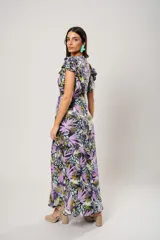 Vestido largo con estampado floral en tonos lila, verde y negro. Tiene escote en V, mangas cortas con volados y corte cruzado con lazo en la cintura.