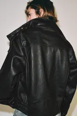 Cazadora bomber de cuero sintético negro, con cuello subido, manga larga, trabillas en los hombros, bolsillos laterales de vivo y bajo elástico. Cierre frontal con cremallera metálica y botones a presión ocultos por solapa.