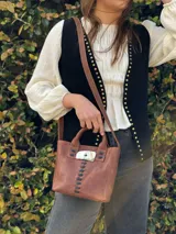 Cartera de cuero marrón de formato estructurado, con doble asa de mano y correa larga ajustable para llevar al hombro o cruzada. Cuenta con detalles de costuras negras a la vista y una pieza decorativa central de resina con efecto carey y aplique metálico.