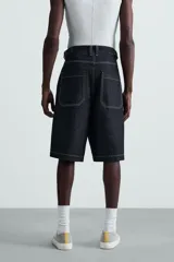 Bermuda de denim rígido color azul oscuro (sin prelavar) con corte amplio (baggy fit). Presenta pliegues frontales, trabillas ajustables en la cintura, bolsillos delanteros y bolsillos traseros de parche, todo resaltado con pespuntes a contraste.