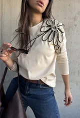 Remera manga larga de color beige, con detalle de aplique floral tridimensional en el hombro confeccionado con cordones en blanco y negro.