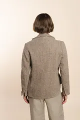 Chaqueta marrón de lana con cuello, solapa y bolsillos ojal. Forro solo en las mangas y terminaciones envivadas. Esquinas redondeadas y mangas con corte en puño que permiten transformarla en manga 3/4. Más larga en la espalda que en la delantera. Abrochadura con dos botones.