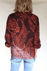 Camisa de lino con estampado paisley en tonos marrón y naranja, cuello con lazos integrados, mangas largas con frunces en hombros y puños elásticos, y cierre frontal con botones de coco.