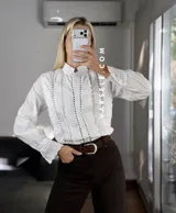 Camisa blanca de estilo romántico con cuello alto, cierre frontal de botones y detalles de encaje y alforzas verticales. Presenta mangas largas con puños decorados con encaje.