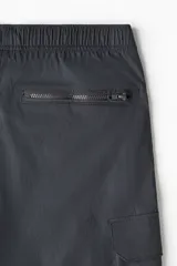 Pantalón cargo negro de nylon, corte recto y holgado, con cintura elástica y cordón ajustable. Presenta bolsillos laterales con cierre, bolsillos cargo con solapa y botones a presión en las piernas, y cordones ajustables en los bajos.