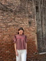 Blusa color chocolate con mangas cortas abullonadas, cuello redondo con volados y detalles de costura en zigzag de color rojo en el pecho y volados oscuros en los hombros.