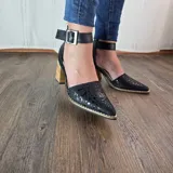 Zapatos de tacón alto negros 100% de cuero, con punta fina, diseño destalonado y tira ajustable al tobillo con hebilla metálica. Tacón ancho de madera y detalle de perforaciones texturizadas en la puntera.