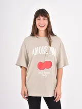 Remera de corte oversize color beige, con cuello redondo y mangas cortas. Presenta un estampado frontal con la frase 'Amore Mio', un dibujo de dos cerezas y la leyenda 'South Tuscany Italy'.