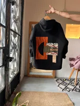 Campera de paño color negro con capucha y diseño de collage en la espalda. El collage incorpora parches en tonos naranja, verde oliva y un tejido con patrón geométrico étnico.