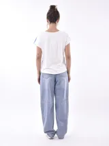 Remera de manga corta, cuello redondo, color blanco con estampado en azul que simula una escena tropical vista desde un interior, con plantas y una figura sentada.