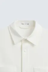 Sobrecamisa negra de tejido con elasticidad, cuello solapa y manga corta. Tiene dos bolsillos de plastrón en el pecho y cierre frontal con botones a presión.